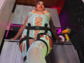 LaurenHills with huge tits, wearing a green lace bodysuit with cutouts, black straps, and fishnet stockings, standing in a room with wooden ceiling and purple lights.