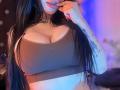 Kkole with big, round tits wearing a ribbed brown crop top, biting her finger with long black hair, a tattoo on her arm, and a necklace.
