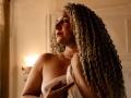 SaraSmiht with long curly blonde hair, pearl necklace, holding a white cloth over her tits, looking to the side, in a warmly lit room, showing a subtle smile.