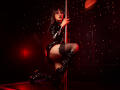TatsumiLee with dark lipstick, wearing a shiny black latex dress, thigh-high boots, and lace top, kneeling by a pole, holding it, showing her bare ass, in a red-lit room.