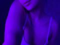 Olieri's upper body in purple-blue light, showing her tits in a white bra, with a tattoo on her neck and her head slightly tilted. Her lips are parted.