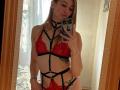 GwynBartell, avec un soutien-gorge en dentelle rouge et de la lingerie noire à bretelles fines, montrant de petites seins et un tatouage sur le côté, prend un selfie devant un miroir avec son téléphone dans une pièce baignée de soleil..