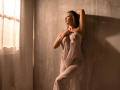 KarlaSoto with her tits covered by a white towel, standing in sunlight, arms raised, looking to the side, her ass slightly visible, in a dimly lit room.