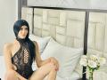 SantiagoDairyFarmer with long black hair, wearing a sheer black lace bodysuit, sitting on a bed with white pillows, legs crossed, showing her pussy through the fabric, smiling.
