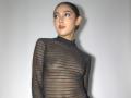 GabySmiith with a bald head, wearing a sheer black dress with horizontal stripes, showing her tits, puffed lips, and earrings, looking to the side, against a plain background.