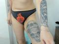 LovesexuellBisou wearing black panties with an orange skull design, showing tattooed arms and thigh with skull tattoos, and a chain bracelet, in a slightly angled photo.