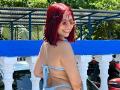 VioletaBlaze with red hair, smiling, wearing a blue bikini, back to camera, showing her ass, tan skin, small tattoos, white-framed glasses, standing by a blue and white railing.