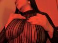 BellaFournierHot with large tits, wearing a sheer black dress with vertical stripes, hands touching chest, red lighting, long dark hair, and hoop earrings, in a sensual pose.