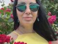 MirandaHott69, com cabelos longos e pretos, óculos de aviador verdes refletivos, brincos azuis, blusa amarela e uma rosa vermelha na mão, mostrando uma pequena tatuagem no pescoço, sorrindo..