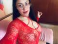 CristalZafiro, con cabello negro largo, gafas rojas y lápiz labial rojo, vestida con una blusa roja de encaje transparente que revela sus pechos, sentada sobre una superficie cubierta de rosa..