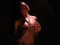 TriciaBarnes with rope bound to her tits, hands near her chest, dim light, dark background, looking up, brown hair, fair skin, small belly button piercing, soft shadows.
