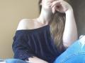 WilsonMelania with long brown hair, off-shoulder black top, blue ripped jeans, and hand near face, looking relaxed and sexy. Her tits are small, and she's wearing a thin necklace.