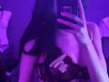 ShylaCatillo taking a selfie with a phone, covering her tits with one hand, wearing a black lace bra, purple-blue lighting, blurred background, long black hair, ring on finger, seductive pose.