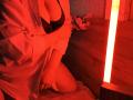 Red-lit photo of Niferttiti with tits in black bra, arms crossed, wearing white shirt, kneeling on bed, hands on thighs, next to glowing rectangular light on wooden wall.