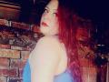 BiancaHottiest with red hair, blue tank top, showing her back and side-boob, looking over her shoulder with red lipstick, in a dimly lit, brick-walled room.