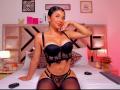AmberSkylerr with tan skin, black lace lingerie, and thigh-high stockings, sitting on a bed, hand near her face, showing her boobs and pussy, smiling confidently.