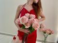 StellaNoir with long red hair in a red satin dress, holding a bouquet of pink roses, wearing red lace lingerie, and a ring, standing next to a glass vase.