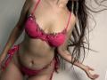 Tisia has pink lace lingerie with floral embroidery, showing her tits and pussy. Her long brown hair is down, and she's leaning slightly forward with her right hand up.