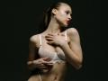 MilagrosEdmisten with her hands on her chest, wearing a white lace bra, showing her tits, and a belly button piercing, against a dark background.