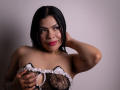 AdrianaPaul with black hair, pink lipstick, and tan skin, wearing a black lace and white frilled strapless top, covering her tits with her hand, smiling.