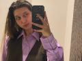 Sanella with long brown hair, wearing a purple shirt and brown vest, taking a mirror selfie with a black phone, showing a neutral expression.