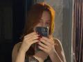 JannaCorsaro with red hair, holding a phone, covering her face, wearing a black lace bra, red nails, mirror selfie, dim light, reflection showing her tits.