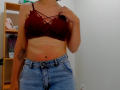 Steisii with tan skin, wearing a red lace bralette and blue denim skirt, holding her right shoulder, showing a tattoo on her left arm.