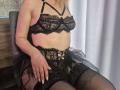Milagres in black lace lingerie with strappy bra, lace garter belt, and sheer black stockings, sitting with hand on thigh, showing her tits and a hint of her ass.