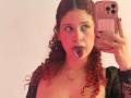 ValeryScot with curly red hair, sticking her tongue out, holding a pink phone, wearing a black lace top, showing her tits, and a necklace against a pink-lit background.