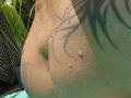 FrenchieLilieFleur's wet ass with water droplets, showing a black tribal tattoo on their tan skin, in a sunlit outdoor setting. Leaf shadows are visible on their butt.