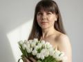 Tilliana, a young woman with straight, brown hair and bangs, holds a bouquet of white tulips. She has light skin and a subtle smile. She's topless, her bare shoulders visible. Her red-painted nails grip the tulips. The white tulips are fresh, with green stems and leaves. Soft light from the left creates a gentle shadow on her face. The image is simple, with a focus on Tilliana and the tulips.