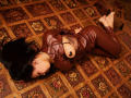 NenaBlack, lying on floral-patterned floor, wearing brown leather bodysuit, exposing black nipple, arms behind back, black hair, tattoo on side, bare feet, brown skin, sexy.