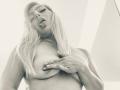 RoxannEmilss tiene el pelo largo y rubio, labios carnosos y está sin sujetador, con una mano cubriendo sus pechos. Está mirando hacia abajo, mostrando su pezón. La foto está en blanco y negro..