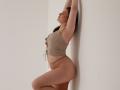 Dreia with arms up, wearing a beige crop top and fishnet panties, leaning against a white wall, showing her ass and a side view of her tits.