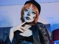 ViolettClay with pale face, black star makeup around eyes, black lipstick, orange bob, black lace robe, choker, finger near mouth, black beret, denim underlay, blue-lit background.