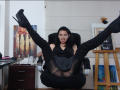 AfroditaRgenis in a black dress, black high heels, and black stockings, legs spread wide, showing her pussy through the sheer fabric, in a chair, looking at the camera.