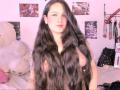 MillaDrew with long, wavy black hair, pale skin, and tattoos, standing topless, showing her small, perky tits, in a pink-lit room with a white teddy bear.