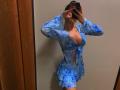 Olieri with blue patterned outfit, revealing cleavage, tied front, ruffled skirt, taking mirror selfie, phone in hand, wood panel on left, beige wall, focused on her tits.