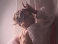 RosaVance with wavy blonde hair, wearing a sheer white blouse, exposing her back and a string of pearls, looking over her shoulder with one hand raised.