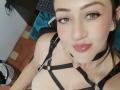 AngelySpencer with light skin, brown eyes, and red lipstick, wearing black strappy harness with silver chains, looking at the camera with a slight pout, in a selfie.
