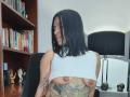 Amelie Blake, met zwart haar, een witte crop top die haar borsten en tatoeages laat zien, zittend in een stoel, naar de zijkant kijkend, met een boekenkast en een lamp op de achtergrond..