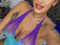 NicolSNaya with tan skin, tattoos, and red lipstick, wearing a purple-blue lace-up halter top, showing her big tits and cleavage, and a pearl necklace, smiling slightly.