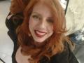 Khharha con i capelli rossi, sorridente, che indossa rossetto rosso, una maglia nera, orecchini dorati, che guarda verso l'alto, seduta, con una bottiglia sullo sfondo, pavimento bianco, capelli leggermente ondulati, pelle chiara..
