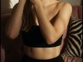 Destinis with light skin, bare shoulders, and black tube top, hands near face, brown hair, and visible ribs, standing in soft light, showing subtle ass and toned midriff.