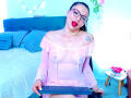 EmelySofia with glasses, red lipstick, and pink off-shoulder dress, showing her small tits, sitting on a chair, holding a keyboard, triangle tattoo on right shoulder.