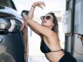 Milagres with tits in a black bikini, leaning on a car, wearing sunglasses, and hoop earrings, touching her head with one hand, at a gas station.