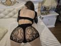TenerliRize, butt in black lace underwear, kneeling on a bed, back to camera, wearing a cutout black top, white tufted headboard, and a white nightstand with a lamp.