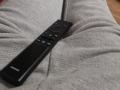 Black Samsung remote control on Tuguinha's gray, textured, woolen ass, with fingers slightly spread. Tuguinha's legs are visible, creating a subtle V-shape.