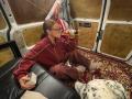 HotCaramelsTrio, wearing a red track jacket and glasses, drinks from a beer can, sitting on a rug, holding a dice, in a dirty, wooden-paneled van.