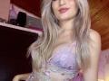 EliseMonet, avec ses longs cheveux blonds, son rouge à lèvres rose et un soutien-gorge en dentelle violette transparent qui dévoile ses seins, est ornée de broderies florales bleues et roses..